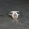 A two-tone ring featuring a pear-cut clear gemstone in a gold bezel setting, mounted on a palladium band, resting on a dark textured slate surface.