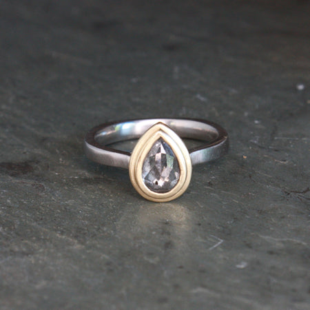 A two-tone ring featuring a pear-cut clear gemstone in a gold bezel setting, mounted on a palladium band, resting on a dark textured slate surface.
