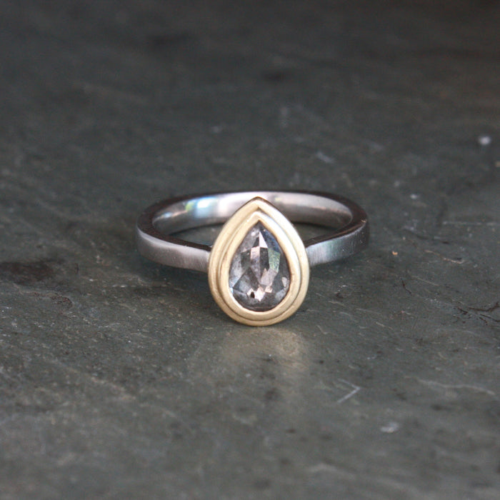 A two-tone ring featuring a pear-cut clear gemstone in a gold bezel setting, mounted on a palladium band, resting on a dark textured slate surface.