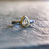 A gold and palladium ring with a pear shaped salt + pepper diamond, resting on a textured slate surface with a blurred background.