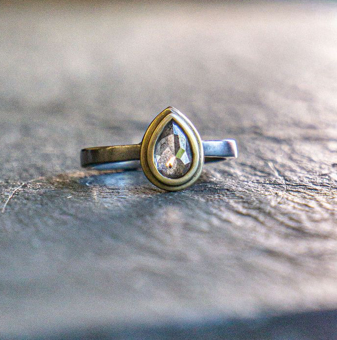 A gold and palladium ring with a pear shaped salt + pepper diamond, resting on a textured slate surface with a blurred background.